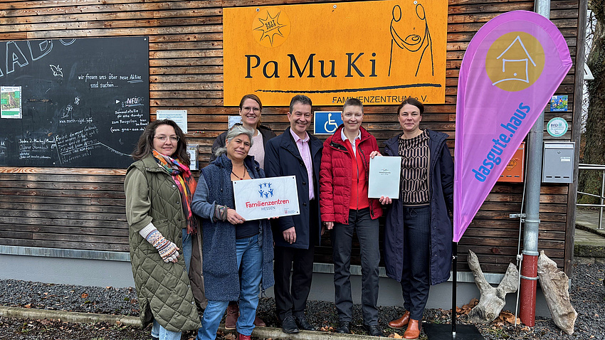 Fünf Frauen und ein Mann stehen vor einer Hauswand. An dieser ist ein Logo zu sehen, ein weiteres findet sich auf einer Beachflag neben der Personengruppe. Eine der Frauen hält eine Tafel mit einem weiteren Logo in Händen, zwei weitere halten gemeinsam ein Dokument hoch.