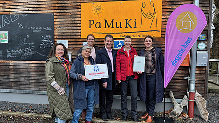 Fünf Frauen und ein Mann stehen vor einer Hauswand. An dieser ist ein Logo zu sehen, ein weiteres findet sich auf einer Beachflag neben der Personengruppe. Eine der Frauen hält eine Tafel mit einem weiteren Logo in Händen, zwei weitere halten gemeinsam ein Dokument hoch.