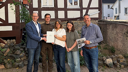 Zwei Frauen und drei Männer stehen gemeinsam vor einem Fachwerkhaus. Eine der Frauen und einer der Männer halten gemeinsam eine Dokumentenmappe in Händen. Ein weiterer Mann hält den Ausdruck einer Internetseite in der Hand.