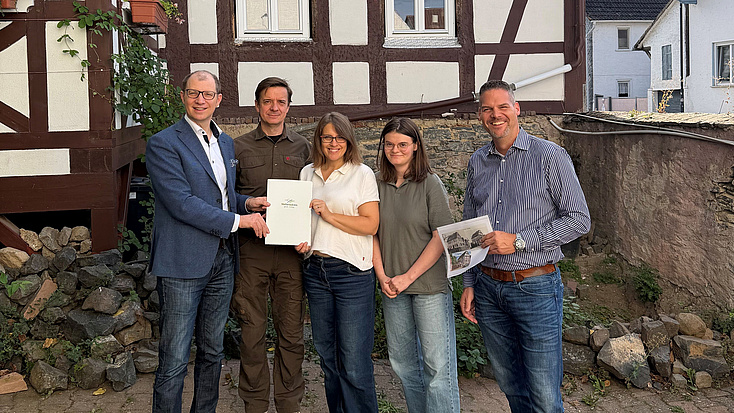 Zwei Frauen und drei Männer stehen gemeinsam vor einem Fachwerkhaus. Eine der Frauen und einer der Männer halten gemeinsam eine Dokumentenmappe in Händen. Ein weiterer Mann hält den Ausdruck einer Internetseite in der Hand.
