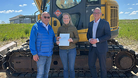 Ein Frau und zwei Männer stehen im Freien vor einem Bagger. Die Frau übergibt einem der Männer ein Dokument.