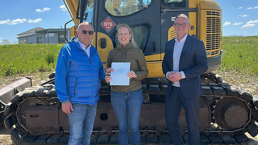 Ein Frau und zwei Männer stehen im Freien vor einem Bagger. Die Frau übergibt einem der Männer ein Dokument.