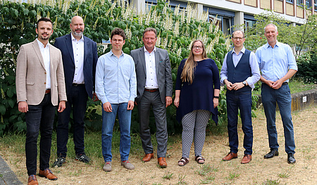 Eine Frau und sechs Männer stehen vor dem Kreishaus. Im Hintergrund  blühende Büsche