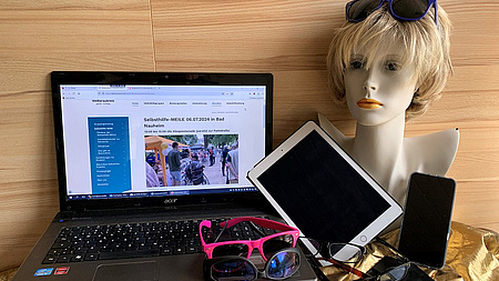 Auf einem Tisch steht ein Laptop. Drumherum liegen mehrere Brillen, ein altes iPad, ein Handy und eine Frauenperücke.