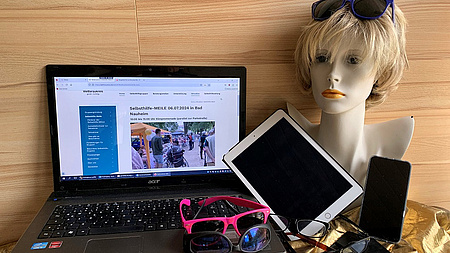 Auf einem Tisch steht ein Laptop. Drumherum liegen mehrere Brillen, ein altes iPad, ein Handy und eine Frauenperücke.