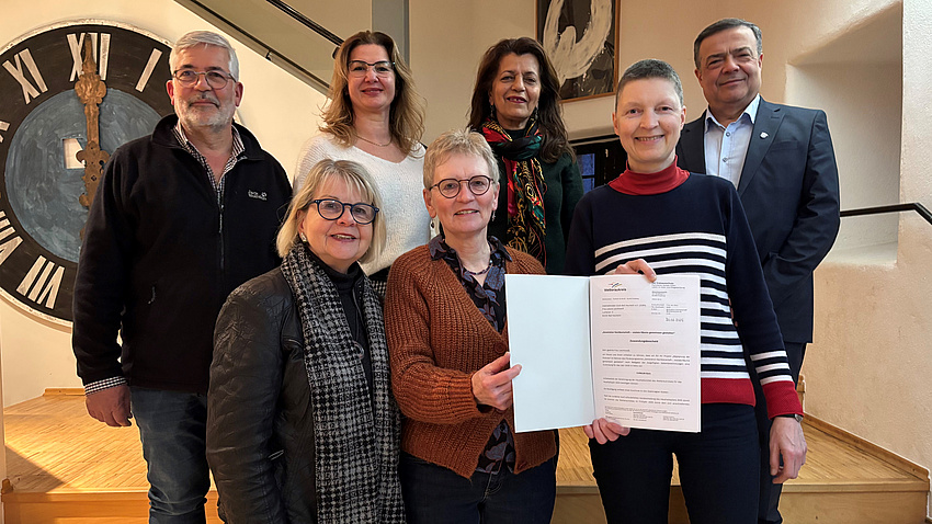 Fünf Frauen und zwei Männer stehen auf einer Treppe. Im Hintergrund ist an der Wand ein großes Ziffernblatt einer Uhr zu sehen. Zwei der Frauen halten gemeinsam ein Dokument hoch.