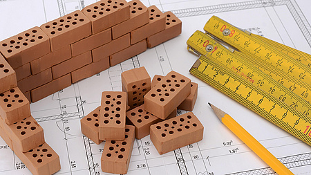 Symbolbild: Ein gezeichneter Plan eines Hauses. Darauf ein Zollstock und Baumaterialien.