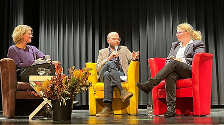 Zwei Frauen und ein Mann auf einer Bühne. Jede Person sitzt auf einem andersfarbigen Sessel. Der Mann in der Mitte spricht in ein Mikrofon.