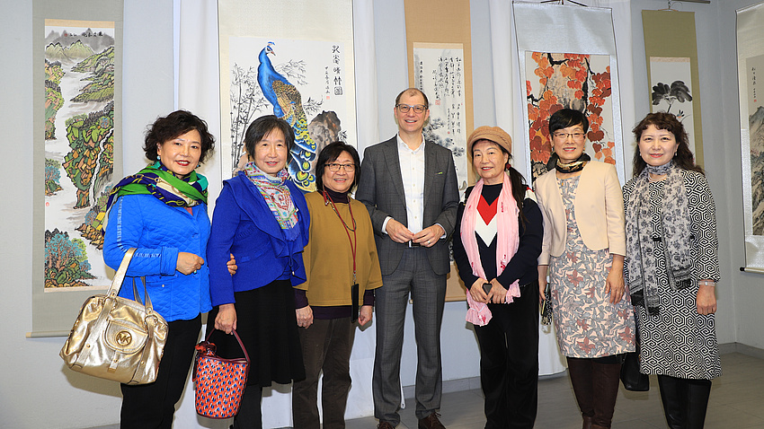 Sechs Frauen und ein Mann stehen in einem Raum vor mehreren Gemälden im chinesischen Stil, die hinter ihnen an der Wand hängen. Diese zeigen Landschafts-, Tier- und Pflanzendarstellungen.