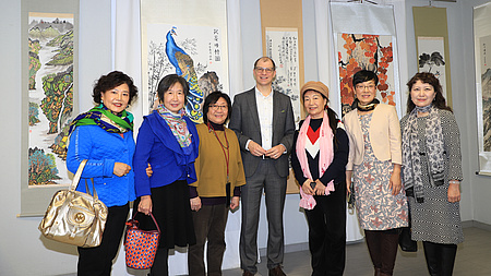 Sechs Frauen und ein Mann stehen in einem Raum vor mehreren Gemälden im chinesischen Stil, die hinter ihnen an der Wand hängen. Diese zeigen Landschafts-, Tier- und Pflanzendarstellungen.