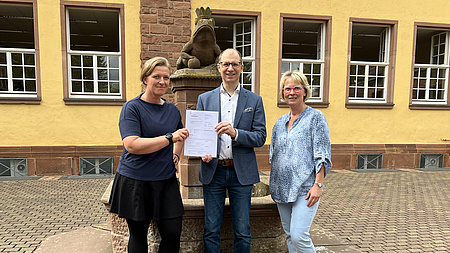 Landrat Jan Weckler, Vereinsvorsitzende Melanie Anschütz und Schulleiterin Sabine Weigandt stehen vor einem Brunnen. Sie halten ein Dokument in der Hand.