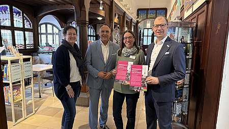 Zwei Frauen und zwei Männer stehen gemeinsam in einem Saal, in dem mehrere Regale mit Büchern stehen. Eine der Frauen und einer der Männer halten zusätzlich Flyer in den Händen.