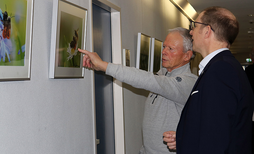 Ein Mann deutet auf eine an der Wand hängende Fotografie und erklärt einem anderen Mann, was auf ihr zu sehen ist.