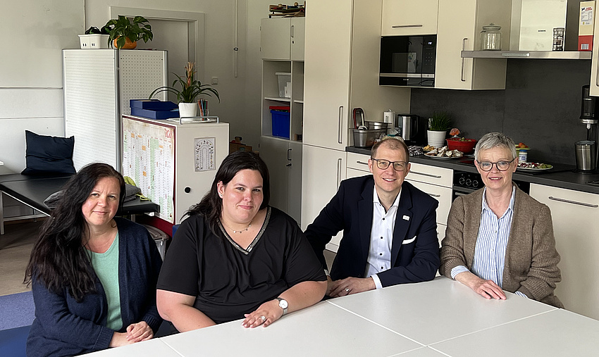 Ein Mann und drei Frauen sitzen an einem Tisch. In Hintergrund eine Küche.