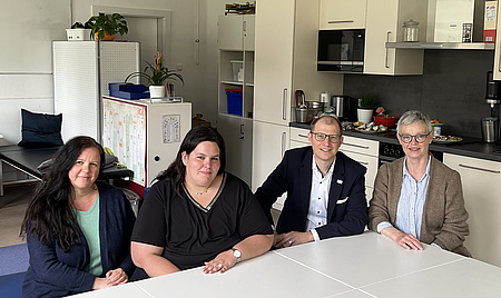 Ein Mann und drei Frauen sitzen an einem Tisch. In Hintergrund eine Küche.