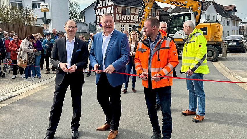 Drei Männer stehen auf einer Straßen und schneiden symbolisch ein Absperrband durch. Im Hintergrund sind weitere Personen ein Bagger und Häuser zu sehen.