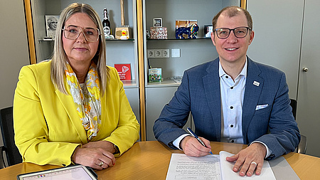 Ein Mann und eine Frau sitzen an einem Tisch. Vor ihnen liegt ein Dokument, das unterschrieben werden muss.