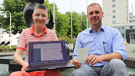 Eine Frau und ein Mann sitzen auf einem Platz im Freien, im Hintergrund ist ein mehrstöckiges Gebäude zu sehen. Die Frau hält ein Laptop in Händen, der Mann zeigt ein Ausweisdokument.