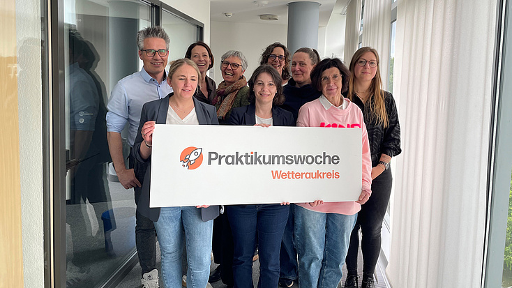 Neun Frauen und Männer stehen in einem Flur. Drei von ihnen halten ein Schild mit einem Logo in Händen.