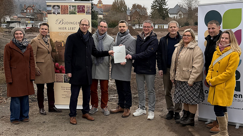 Vier Frauen und sechs Männer stehen gemeinsam im Freien. Im Hintergrund ist ein Baustellengelände zu sehen. Vier der Männer in der Bildmitte halten gemeinsam ein Dokument in Händen.