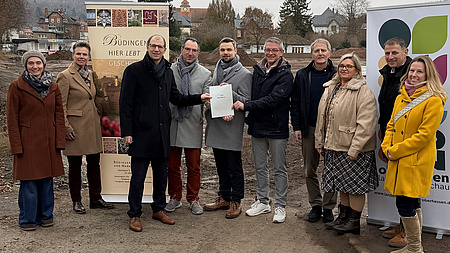 Vier Frauen und sechs Männer stehen gemeinsam im Freien. Im Hintergrund ist ein Baustellengelände zu sehen. Vier der Männer in der Bildmitte halten gemeinsam ein Dokument in Händen.