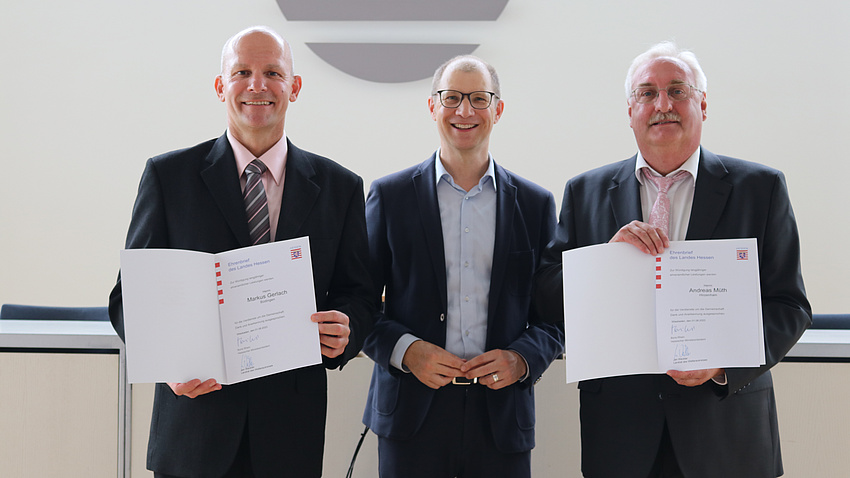Drei Männer stehen nebeneinander und lächeln in die Kamera. Zwei von ihnen halten eine Urkunde in der Hand.