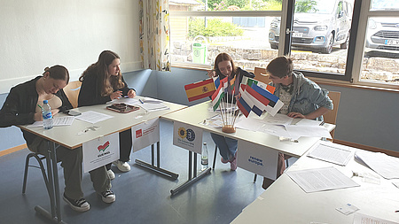 Vier Jugendliche sitzen an einem Tisch, auf dem verschiedene Flaggen der EU aufgestellt sind, vor ihnen hängen Schilder verschiedener europäischer Parteien