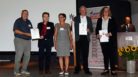 Eine kleine Personengruppe steht auf einer Bühne. Sie lächeln in die Kamera und haben Urkunden in der Hand.