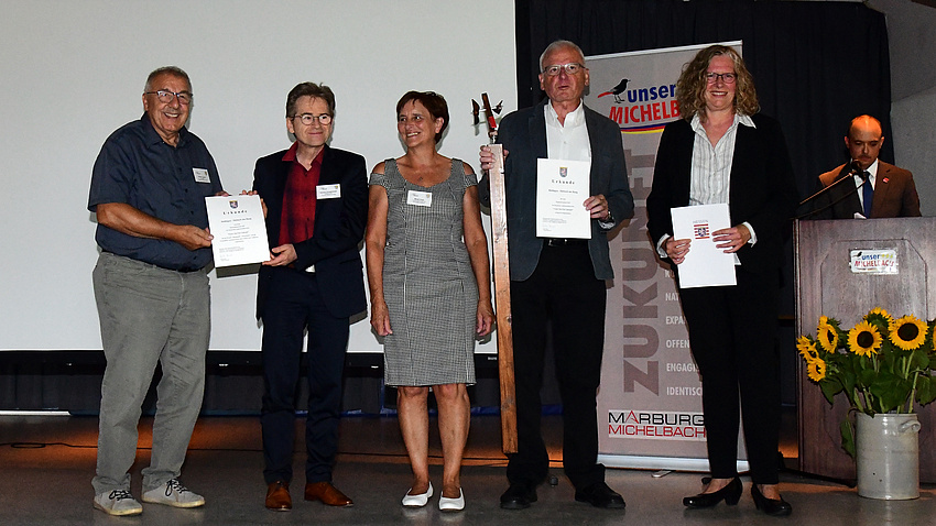 Eine kleine Personengruppe steht auf einer Bühne. Sie lächeln in die Kamera und haben Urkunden in der Hand.