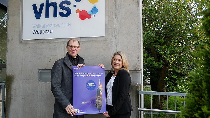Ein Mann und eine Frau halten ein lilafarbenes Plakat in die Höhe. Auf dem Gebäude im Hintergrund hängt ein Schild "vhs".