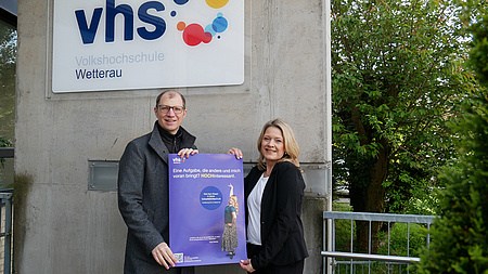 Ein Mann und eine Frau halten ein lilafarbenes Plakat in die Höhe. Auf dem Gebäude im Hintergrund hängt ein Schild "vhs".