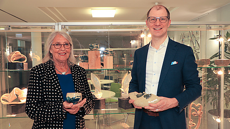 Eine Frau und ein Mann stehen in einem größeren Raum vor einer Vitrine, in der sich Keramik-Kunstwerke befinden. Beide halten je ein Keramik-Kunstwerk in Händen.