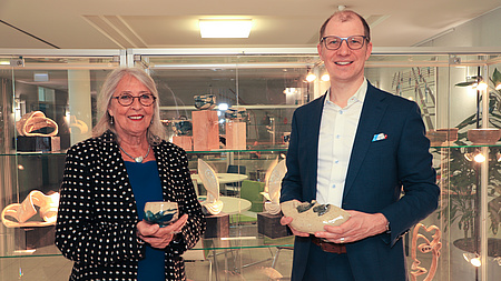 Eine Frau und ein Mann stehen in einem größeren Raum vor einer Vitrine, in der sich Keramik-Kunstwerke befinden. Beide halten je ein Keramik-Kunstwerk in Händen.