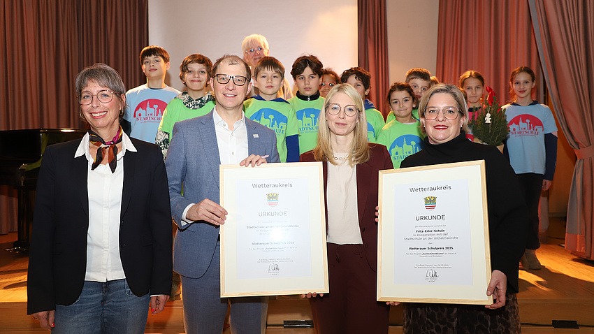 Auf und vor der Bühne eines Veranstaltungssaals stehen zahlreiche Kinder, drei Frauen und ein Mann. Der Mann und zwei der Frauen halten zusammen zwei gerahmte Urkunden in Händen.