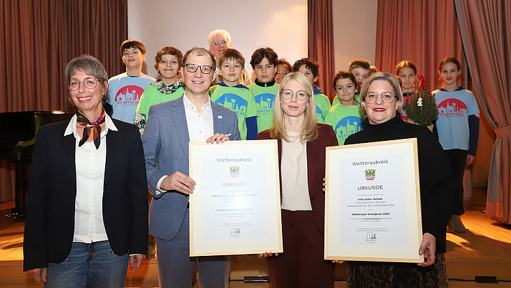 Auf und vor der Bühne eines Veranstaltungssaals stehen zahlreiche Kinder, drei Frauen und ein Mann. Der Mann und zwei der Frauen halten zusammen zwei gerahmte Urkunden in Händen.