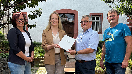 Vier Menschen stehen auf einem Kopfsteinpflasterhof und halten eine Urkunde in die Kamera.