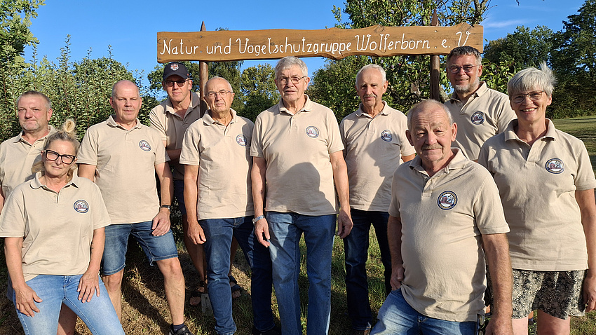 Eine Gruppe von Menschen steht vor einem Holzschild mit der Aufschrift Natur- und Vogelschutzgruppe Wolferborn e.V.