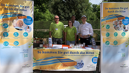 Eine Frau und drei Männer stehen in einem Parkgelände an einem Stand, der links und rechts von je einem Banner eingerahmt ist.