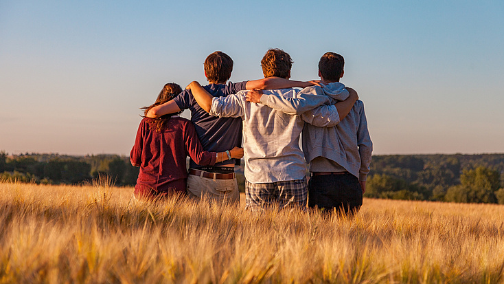 Wir sehen vier junge Menschen in einem Kornfeld, in einer Reihe stehend, eng miteinander verbunden. Arm in Arm. Sie blicken weit in die Ferne.