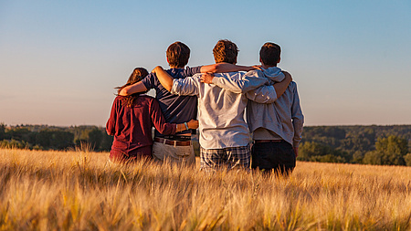 Wir sehen vier junge Menschen in einem Kornfeld, in einer Reihe stehend, eng miteinander verbunden. Arm in Arm. Sie blicken weit in die Ferne.
