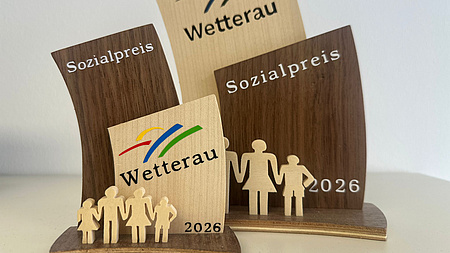 Zwei aus Holz gestaltete Skulpturen stehen nebeneinander. Stilisiert ist auf ihnen eine vierköpfige Familie abgebildet, bestehend aus den Eltern und einem Jungen sowie einem Mädchen. Beide Skulpturen sind mit den Worten "Sozialpreis Wetterau 2026" beschriftet.