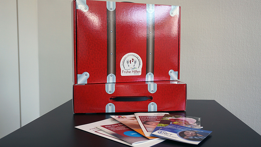 Ein kleiner roter Koffer aus Pappe. Darauf ein Aufkleber mit dem Logo der Frühen Hilfen. Vor dem Koffer liegen einige Broschüren