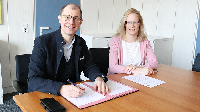 Ein Mann und eine Frau sitzen in einem Büro an einem Tisch. Vor ihnen auf dem Tisch liegt ein Dokument, das der Mann gerade unterschreibt.