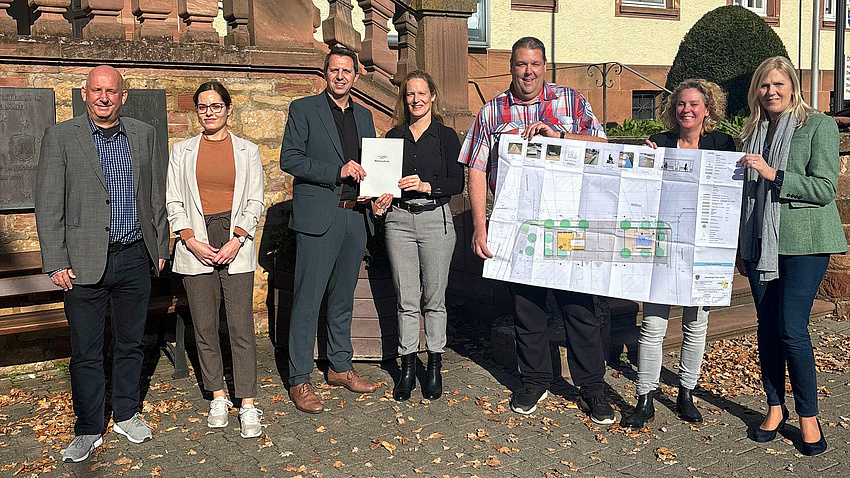Eine Gruppe von Leuten vor einem Gebäude. Sie halten einen großen Bauplan in der Hand.