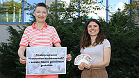 Zwei Frauen stehen im Freien vor einem Gebäude. Eine der Frauen hält ein Plakat in Händen, die andere kleine Informationskarten.