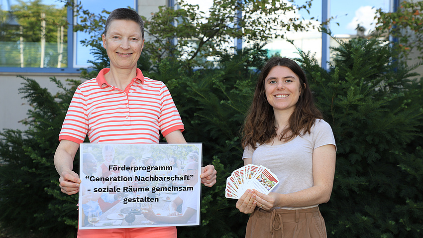 Zwei Frauen stehen im Freien vor einem Gebäude. Eine der Frauen hält ein Plakat in Händen, die andere kleine Informationskarten.