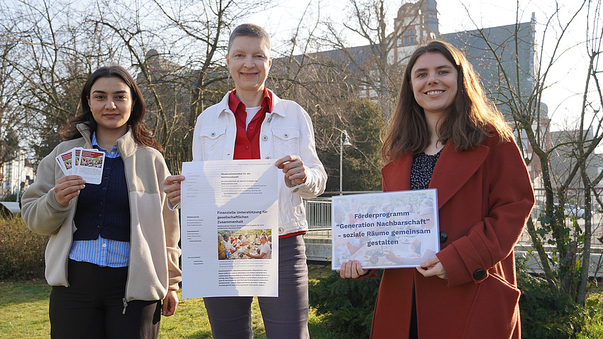 Drei Frauen stehen im Freien auf einem Platz, im Hintergrund sind Büsche und Gebäude zu sehen. Zwei der Frauen halten Plakate in Hände, die dritte mehrere Karten.
