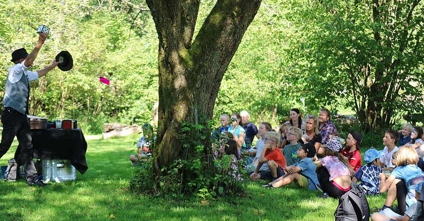 Kinder und Erwachsene sitzen auf einer Wiese. Im Vordergrund ein Baum. Alle sehen einem Mann zu, der Zauberkunststücke vorführt.