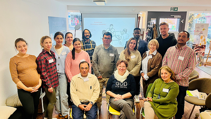 15 Frauen und Männer stehen und sitzen in einem Raum beieinander.