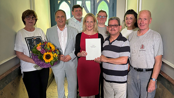 Acht Personen, vier Frauen und vier Männer, stehen im Flur eines Hauses. Eine der Frauen übergibt eine Urkunde an einen der Männer. Eine weitere Frau hält einen Blumenstrauß in der Hand.